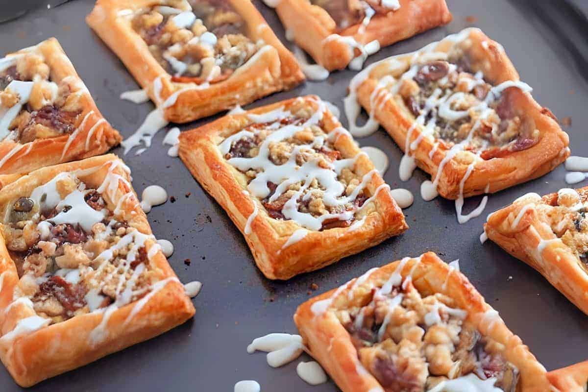 Golden brown Dutch Apple Danishes, topped with a fruit and nut filling, drizzled with white icing, displayed on a dark baking sheet.