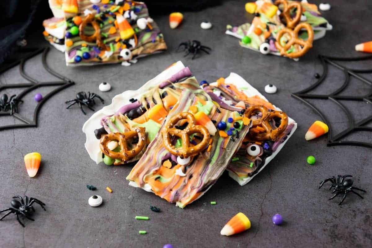 Halloween Candy bark topped with pretzels, candy eyes, sprinkles, and candy corn, displayed on a dark surface with toy spiders and web decorations.