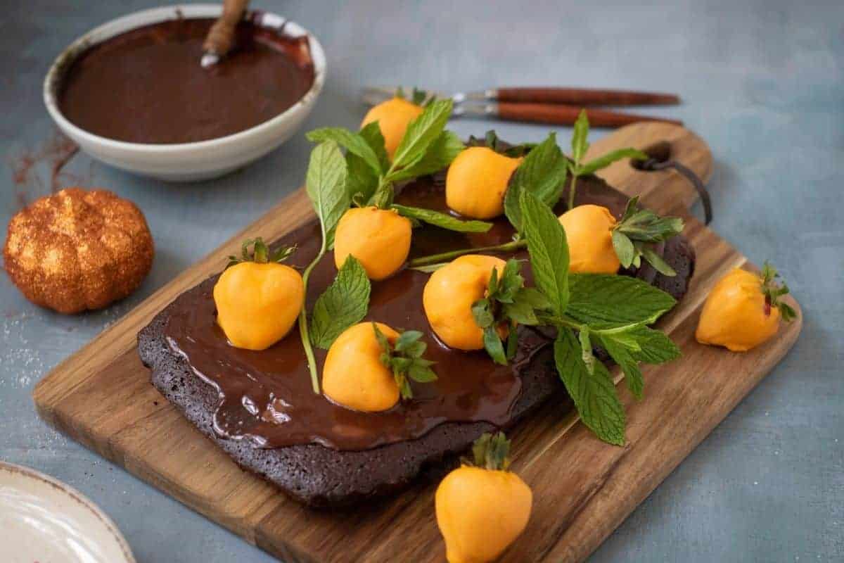 A rectangular Halloween Pumpkin Patch Brownie Cake, topped with yellow fruit resembling mini pumpkins and fresh mint, sits on a wooden board.