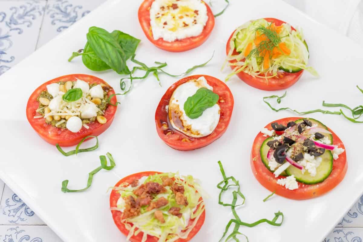 Six tomato slices topped with various ingredients, including cheese, lettuce, olives, herbs, and garnishes, arranged on a white plate with basil leaves and shredded greens.