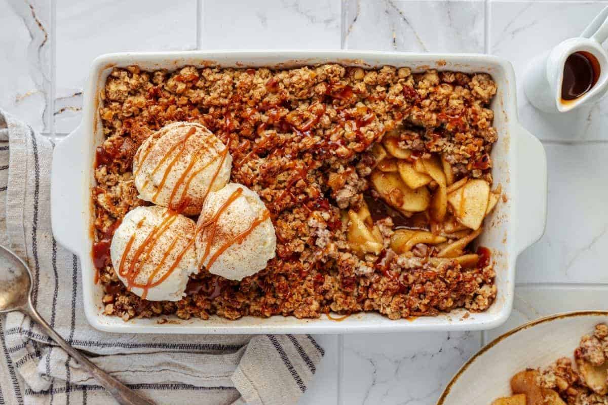 An Apple Crisp with Vanilla Ice Cream, served in a rectangular dish, topped with two scoops of vanilla ice cream and a caramel drizzle, accompanied by a portion on a plate beside it.