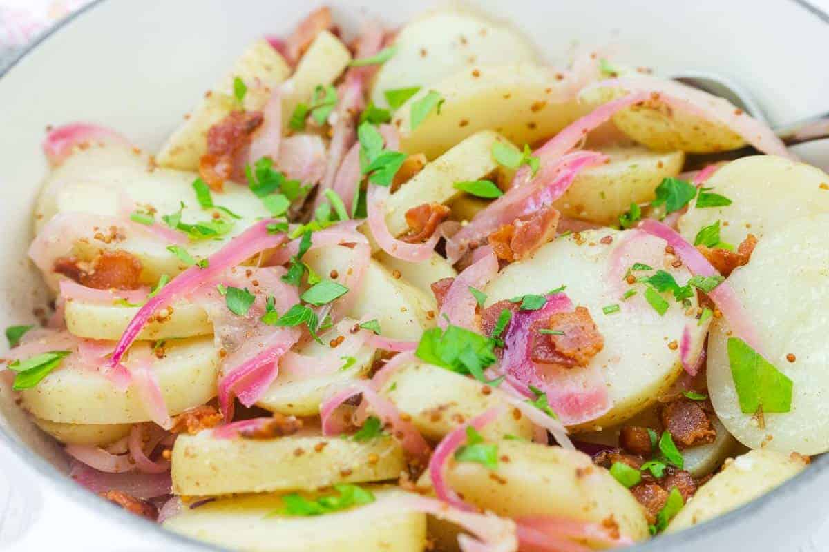 Chef Jenn’s Warm German Potato Salad with sliced potatoes with bacon, red onions, and chopped parsley, served in a white bowl.