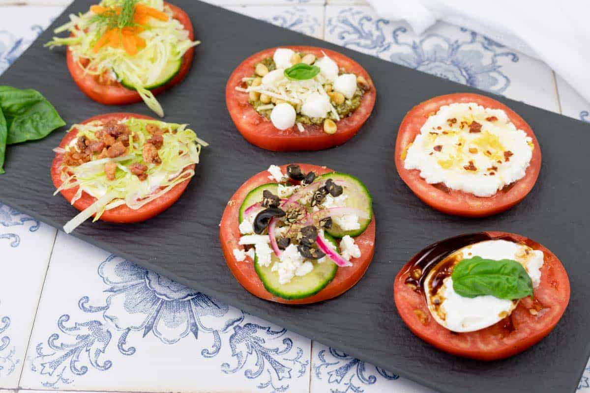 Six pieces of tomatoes with different toppings on a black slate plate.