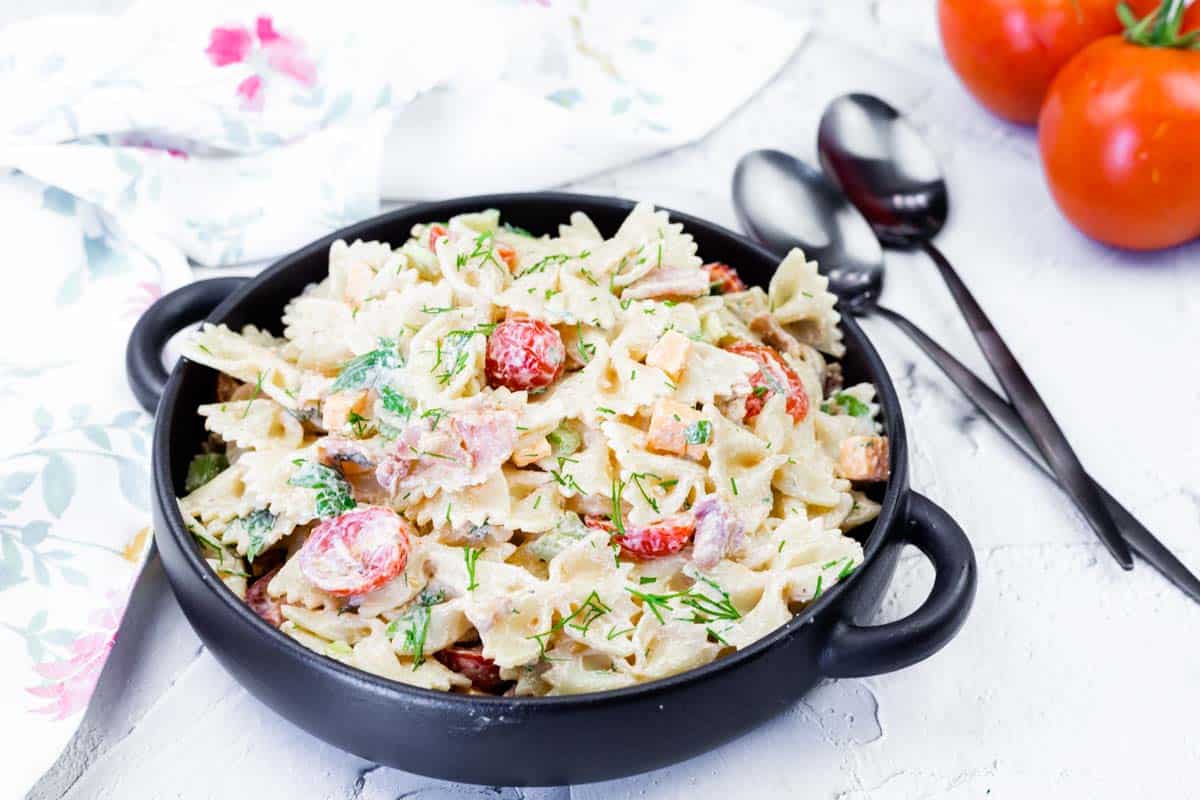 A black bowl filled with picnic pasta salad with chopped vegetables and herbs, placed on a white surface next to spoons and fresh tomatoes.