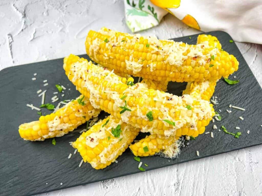 Several pieces of Parmesan Garlic Corn Ribs, topped with grated cheese and chopped herbs, are arranged on a black slate serving board.