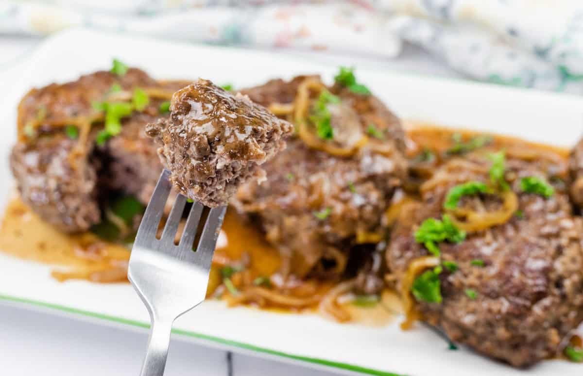 A close-up of a fork holding a piece of cooked Mississippi Salisbury steak with gravy and onions, with more steak pieces and sauce on a white plate in the background.