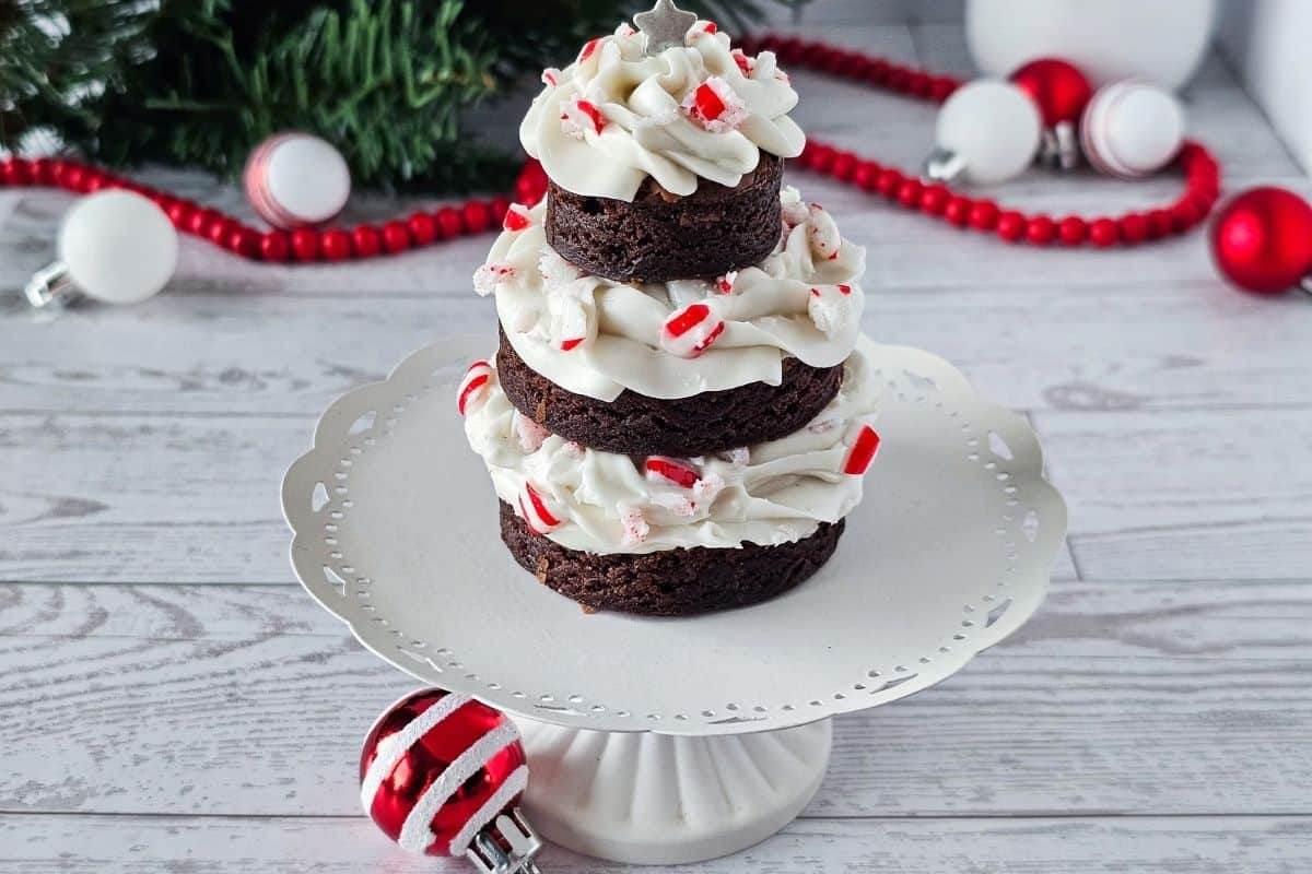 A stack of three round chocolate brownies with white frosting and crushed peppermint, arranged in a tree shape on a white cake stand with holiday decorations in the background.