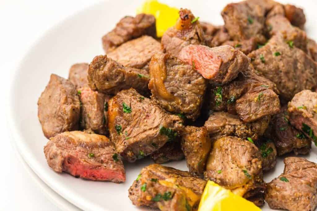 A plate of cooked steak bites garnished with herbs, served with pieces of yellow bell pepper.