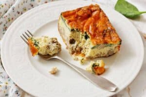 A slice of Sausage, Spinach, and egg casserole on a white plate, with a fork holding a bite-sized piece.