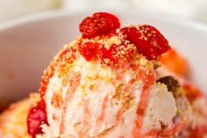 A close-up of a bowl of ice cream topped with strawberry pieces, strawberry sauce, and crumbled biscuit or cookie crumbs.