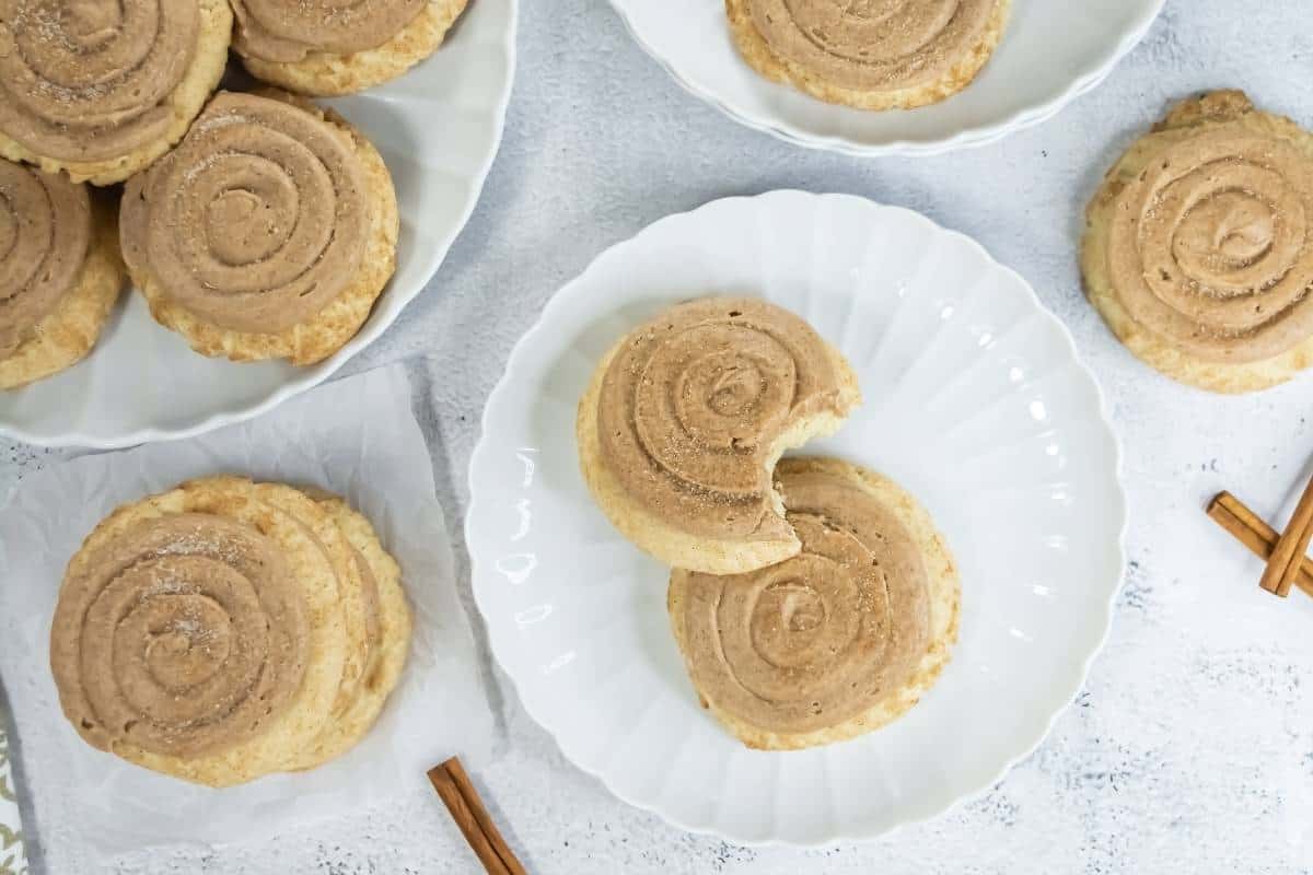Crumbl Copycat Churro Cookies are arranged on white plates and a napkin. One cookie on a plate has a bite taken out. Two cinnamon sticks are placed nearby on the white surface.