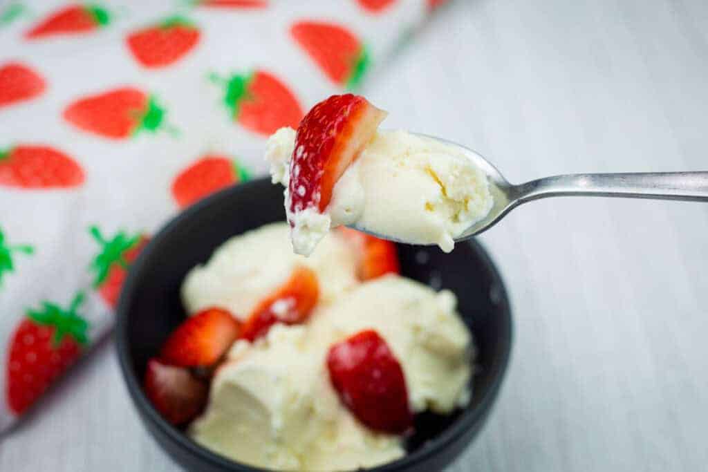 A spoonful of vanilla ice cream and a strawberry held above a bowl with more ice cream and strawberries, next to a strawberry-patterned cloth.