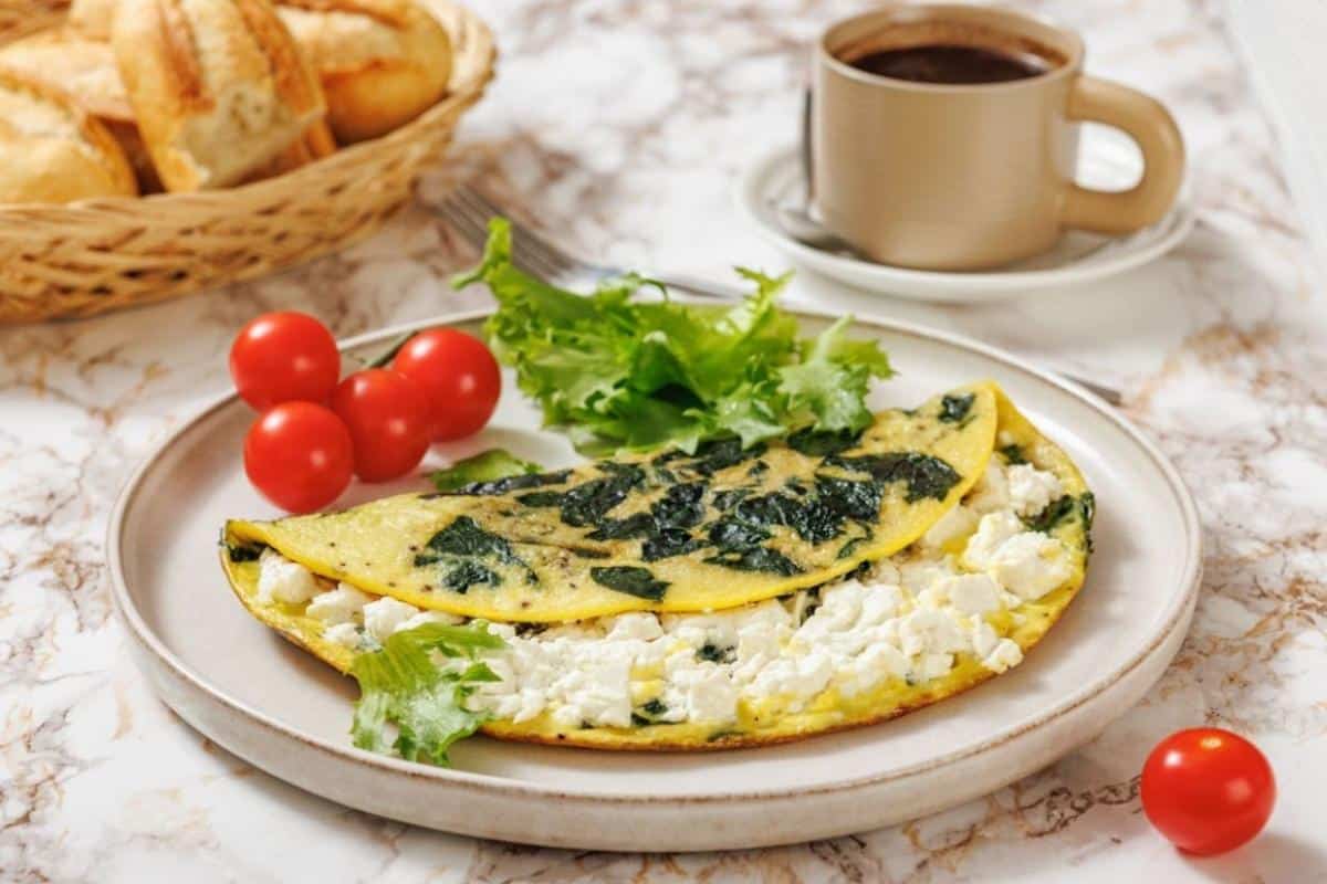 A plate with a spinach and goat cheese omelette, fresh lettuce, and cherry tomatoes, next to a cup of coffee and a basket of bread rolls on a marble table.