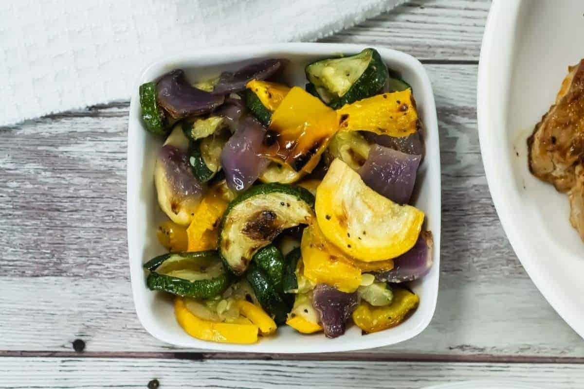 A white bowl filled with broiled vegetables, including zucchini, yellow squash, and red onion, sits on a wooden surface next to a white towel.