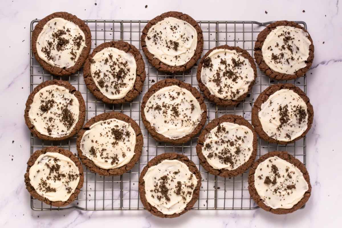 A cooling rack with twelve Crumbl Copycat Frosted Oreo Cookies topped with white frosting and chocolate crumbs, arranged on a white marble surface.