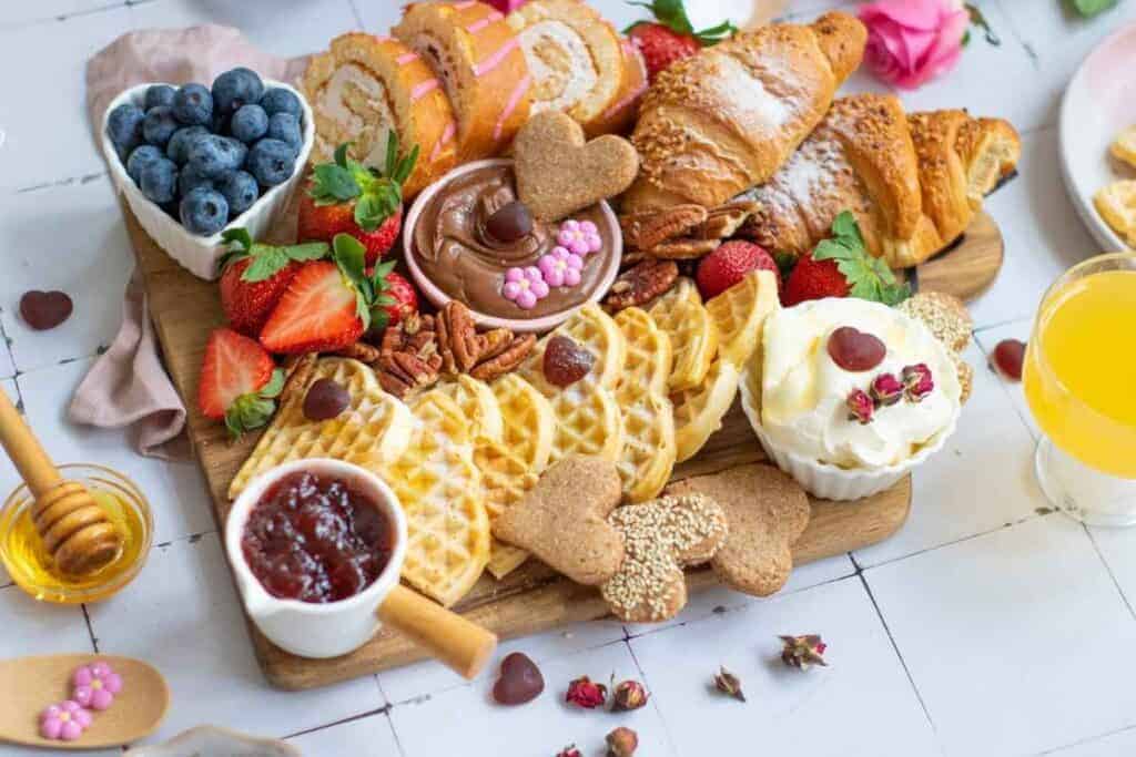 A Valentine's breakfast platter with waffles, croissants, strawberries, blueberries, jam, whipped cream, chocolate spread, cookies, and orange juice on a wooden board.