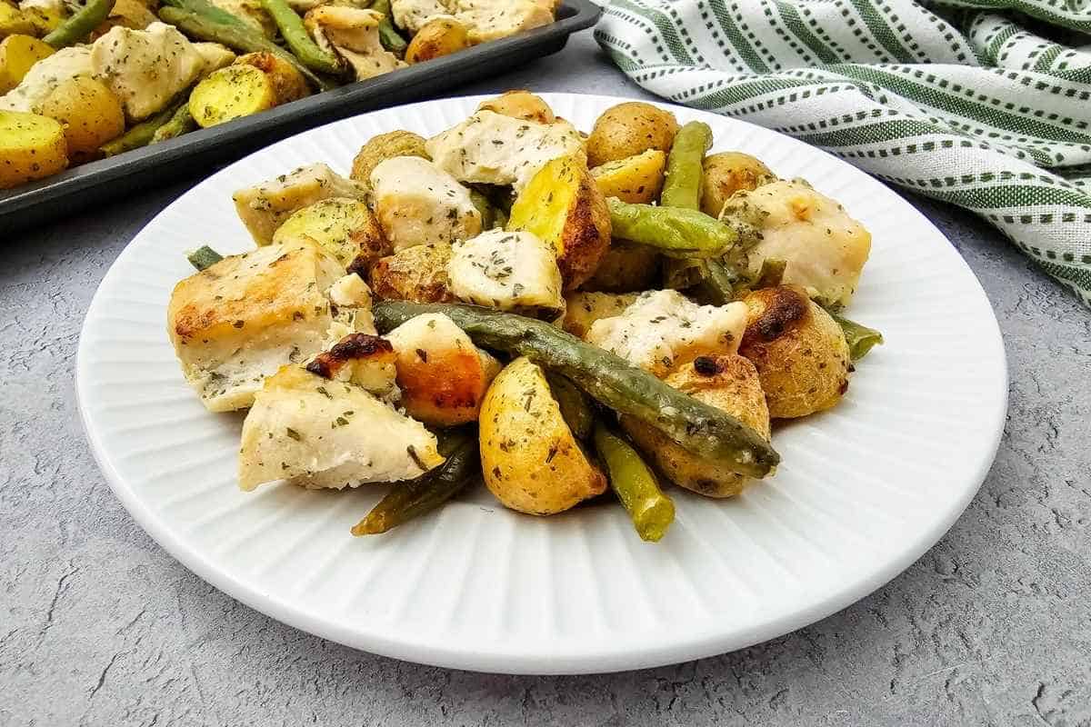 A white plate with roasted chicken pieces, halved baby potatoes, and green beans, with a baking tray and striped towel in the background.