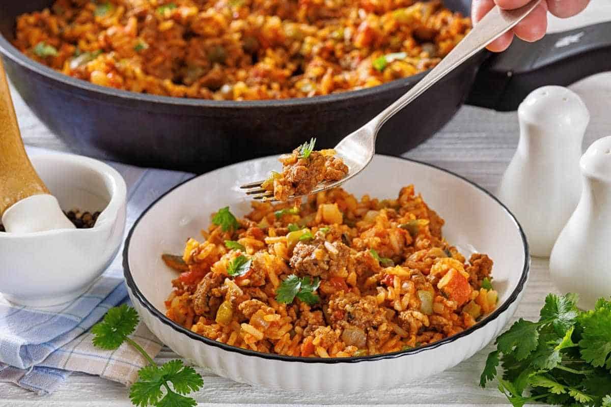 A bowl of Texas hash with rice garnished with cilantro, with a fork lifting a bite, and a skillet of the same dish in the background.