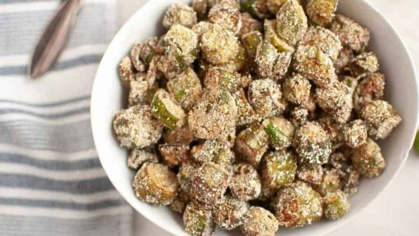 A white bowl filled with crispy, breaded and fried okra pieces, placed on a light surface next to a striped cloth napkin.