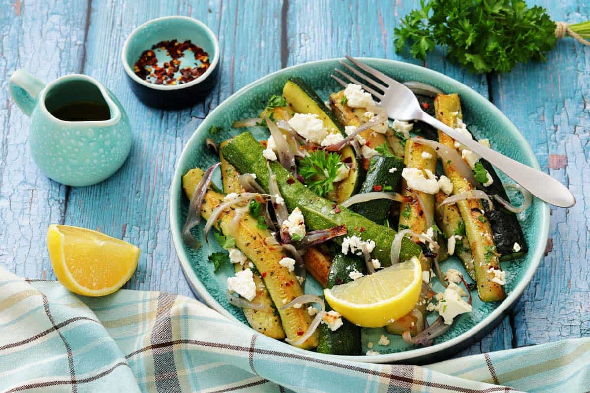 A plate of roasted zucchini with onions, crumbled cheese, lemon wedges, and herbs on a blue wooden table, accompanied by a fork, napkin, and small dishes of oil and chili flakes.