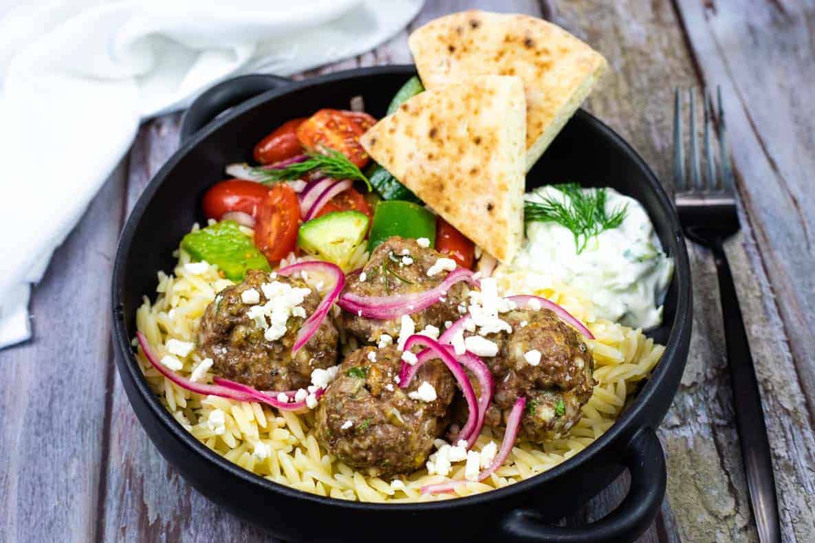 A black bowl with orzo pasta, meatballs topped with feta and pickled onions, Greek salad, pita bread, and a side of tzatziki sauce on a rustic wooden table.