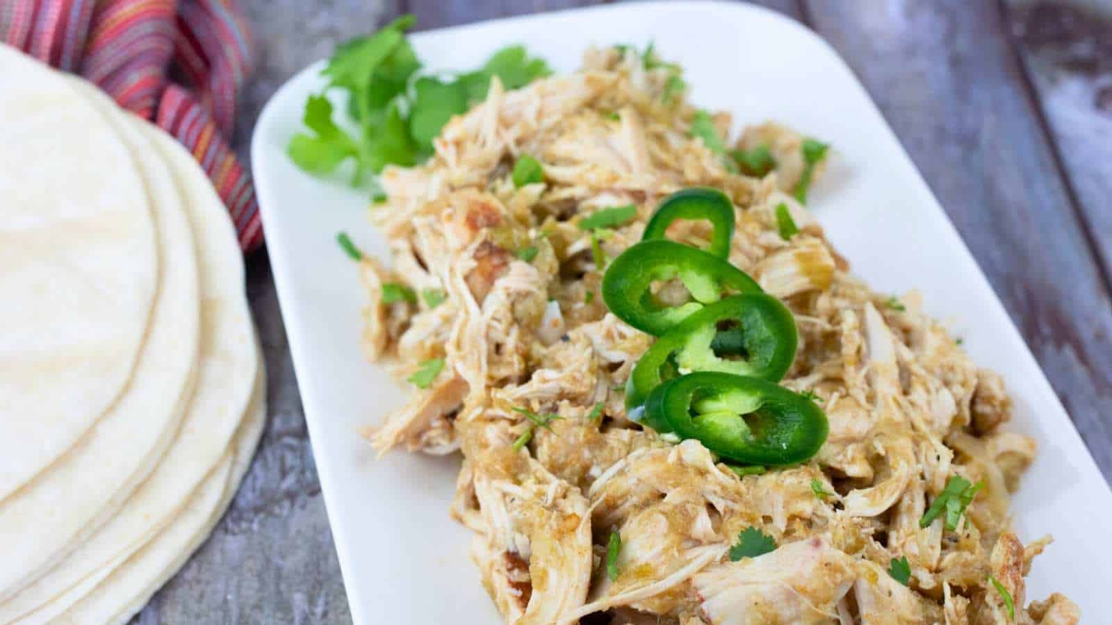 Slow Cooker Hatch Chili Salsa Chicken topped with sliced jalape&ntilde;os and cilantro on a white plate, next to a stack of tortillas.